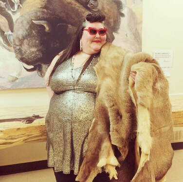 Shilo George is a light brown skinned person, who in this photo is wearing a metallic gold shirt, red sunglasses, and a fur pashmina. Shilo is striking a sassy pose.