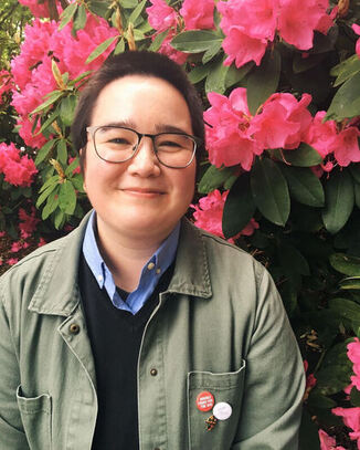Steph Ng Ping Cheung is a light brown skinned person with brown glasses and a dark brown buzzcut. They are smiling without teeth in this photo; they are wearing a green jacket with a dark blue sweater and a light blue collared shirt, and are standing in front of pink flowers.