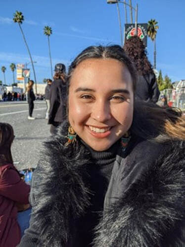 Lucinda Law is a medium brown skinned person wearing a black jacket with a faux fur hood in this photo. She has long black hair, dark brown eyes, and she's standing outside in a crowded area.