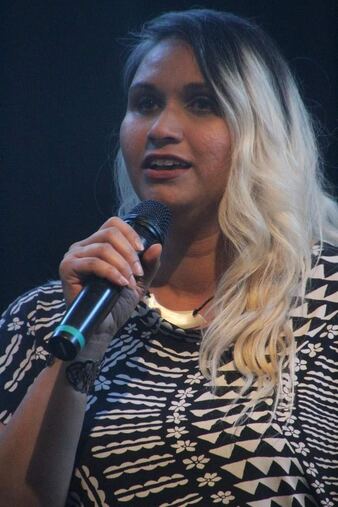 Virginia Luka is a medium brown skinned person with dark brown eyes, and blond hair with dark roots in this photo. Virginia is wearing a geometric black and white shirt and is speaking into a microphone.